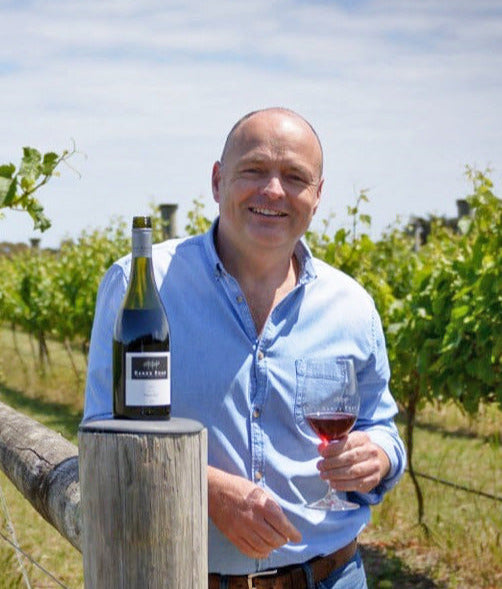 Will Durham holding a glass of red wine and a bottle in the middle of a lush vineyard at Banks Road.